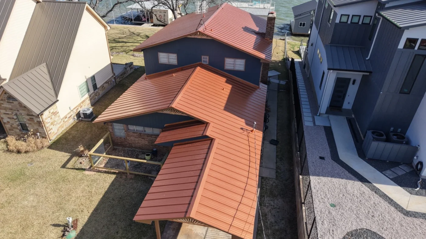 Aerial view of a lakeside home with a red metal roof surrounded by neighboring houses.